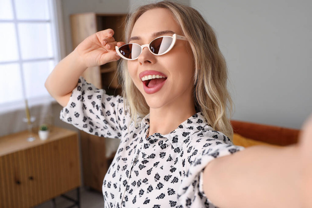 Young woman in stylish sunglasses taking selfie at home, closeup - Foto, afbeelding