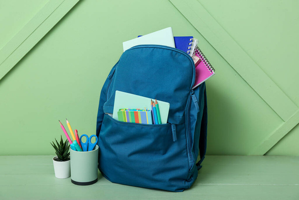 Blue school backpack with notebooks, pencils and houseplant on wooden table near green wall - Foto, afbeelding