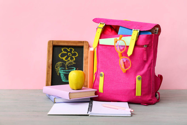 Рюкзак розовой школы с книгами, мелом рисунок цветок и яблоко на сером деревянном столе рядом с цветной стеной - Фото, изображение