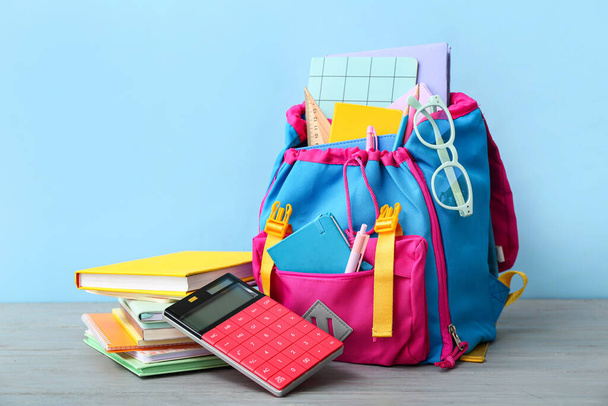 Plecak szkolny z kalkulatorem, książkami i okularami na szarym drewnianym stole przy niebieskiej ścianie - Zdjęcie, obraz