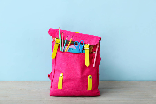 Ροζ σχολική τσάντα πλάτης με ψαλίδι, σημειωματάρια και μολύβια σε γκρι ξύλινο τραπέζι κοντά σε μπλε τοίχο - Φωτογραφία, εικόνα