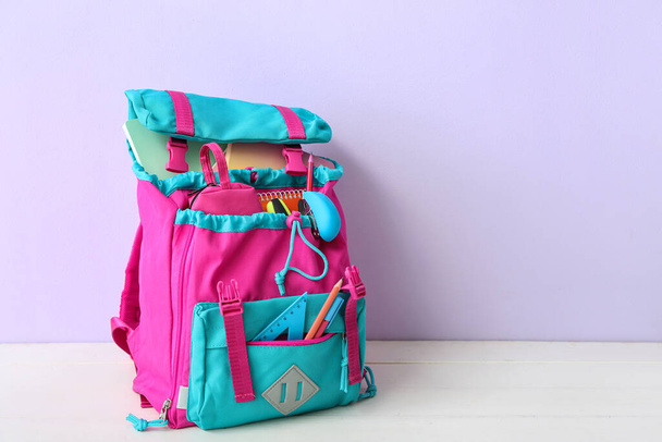 Renkli okul çantası, içinde not defterleri, kalem kutusu ve zımba var. Beyaz ahşap masa üzerinde, leylak duvarın yanında. - Fotoğraf, Görsel