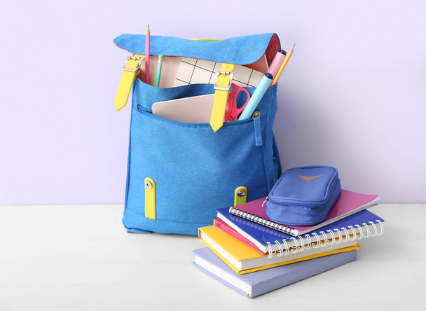 Μπλε σχολική τσάντα πλάτης με βιβλία, κασετίνα και μαρκαδόρους σε λευκό ξύλινο τραπέζι κοντά σε λιλά τοίχο - Φωτογραφία, εικόνα