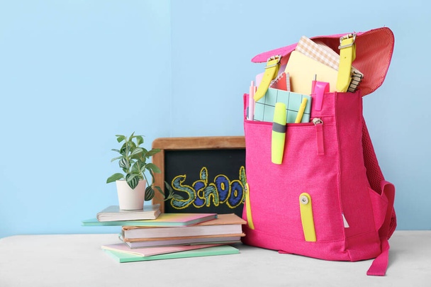 Ροζ σχολική τσάντα πλάτης με σημειωματάρια, μικρό chalkboard και houseplant σε λευκό grunge τραπέζι κοντά στο μπλε τοίχο - Φωτογραφία, εικόνα