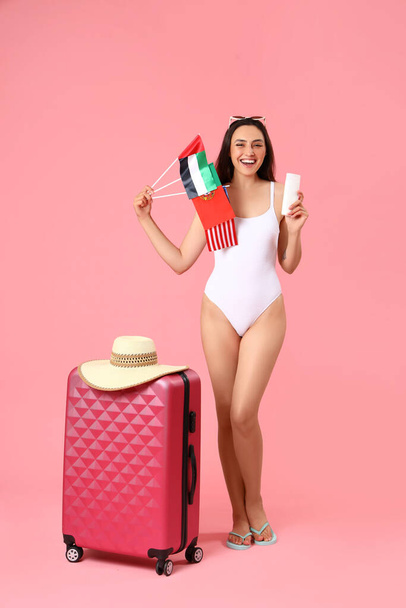 Young woman with sunscreen cream, flags and suitcase on pink background - Foto, Bild
