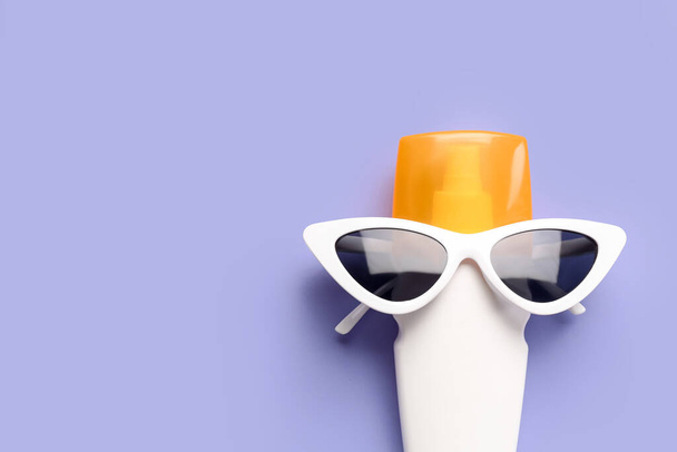 Creatieve compositie met zonnebril en fles zonnebrandcrème op lila achtergrond - Foto, afbeelding