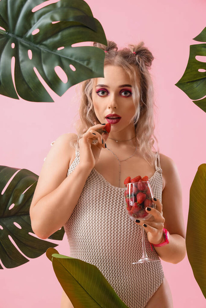 Young woman in swimsuit with glass of strawberries and palm leaves on pink background - Foto, Imagen