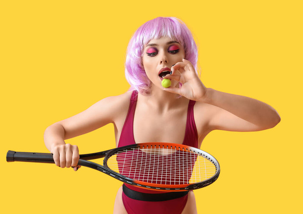 Mujer joven en peluca con dulces y raqueta de tenis sobre fondo amarillo - Foto, imagen