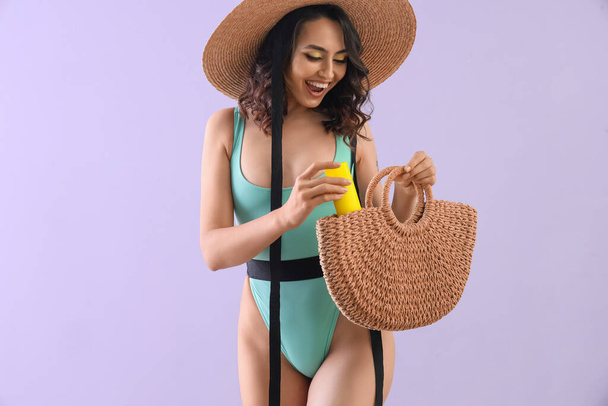 Junge Frau mit Sonnencreme in Strandtasche auf fliederfarbenem Hintergrund - Foto, Bild