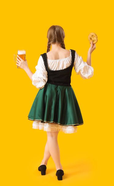 Gyönyörű Octoberfest pincérnő sörrel és pereccel sárga háttérrel, visszapillantás - Fotó, kép