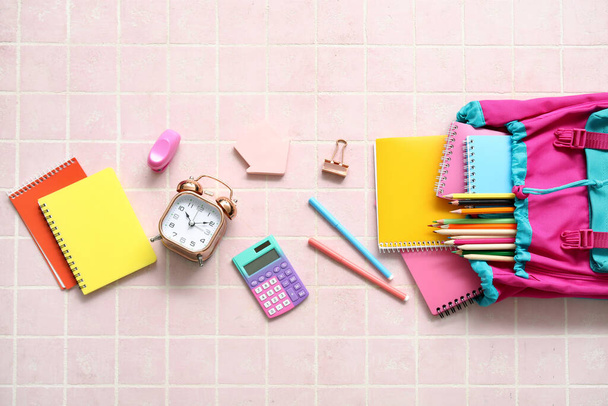 Барвистий шкільний рюкзак з блокнотами, будильником та олівцями на фоні рожевої плитки - Фото, зображення