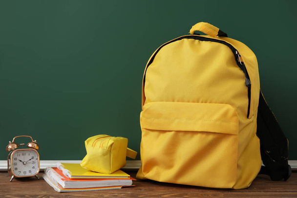 Mochila escolar amarela com despertador, estojo de lápis e cadernos em mesa de madeira perto de quadro-negro verde - Foto, Imagem