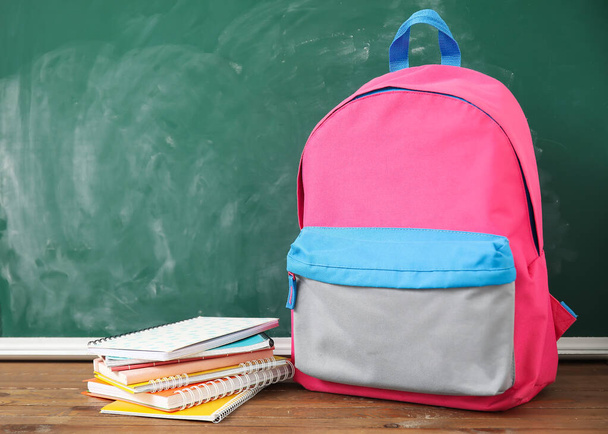 Ροζ σχολική τσάντα πλάτης με σημειωματάρια σε ξύλινο τραπέζι κοντά σε πράσινο πίνακα - Φωτογραφία, εικόνα