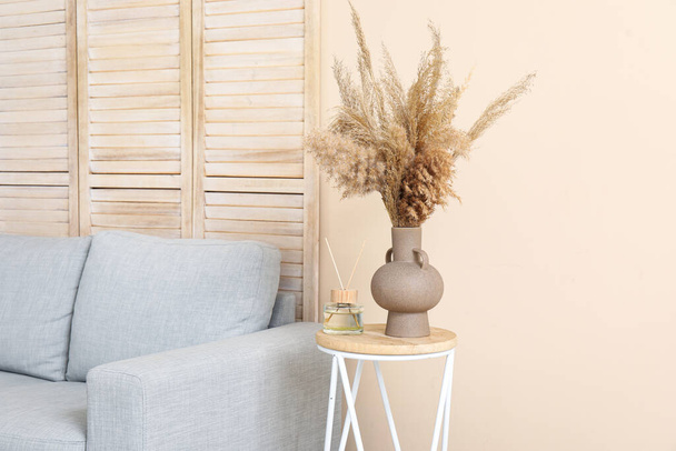 Table with aroma diffuser, pampas grass and sofa near beige wall in room - Valokuva, kuva
