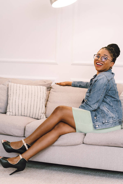 African American woman in light green dress with jacket sitting on sofa, Happy black girl in living room - Foto, afbeelding