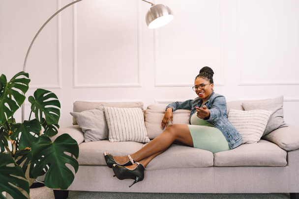 African American woman in light green dress with jacket sitting on sofa, Happy black girl in living room - Фото, изображение