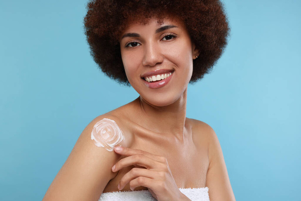 Beautiful young woman applying body cream onto shoulder on light blue background - Фото, зображення