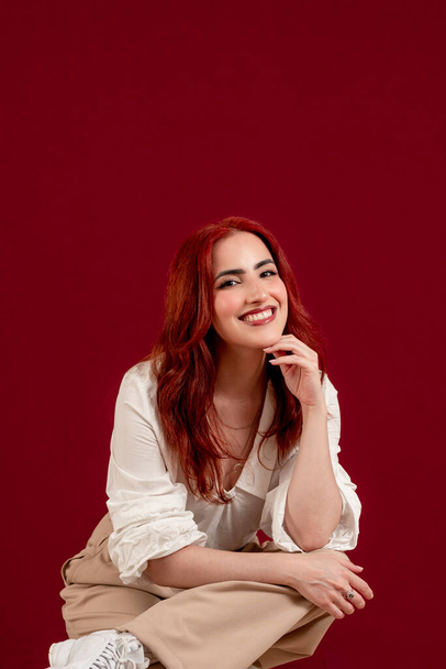 Stylish red-haired woman looking at the camera while posing against a red isolated background. - Photo, Image