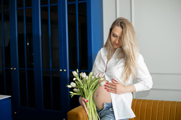 Roztomilá těhotná žena v bílé košili a modré džíny stojící před žlutou pohovkou v bytě s kyticí tulipánů v ruce. Dáma dívá na holé bříško, hladí břicho a jemně se usmívá. - Fotografie, Obrázek