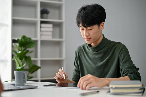 Egy koncentrált és okos ázsiai férfi alkalmi ruhákban, digitális tabletjét használva, hogy az irodai asztalánál intézze a dolgait.. - Fotó, kép