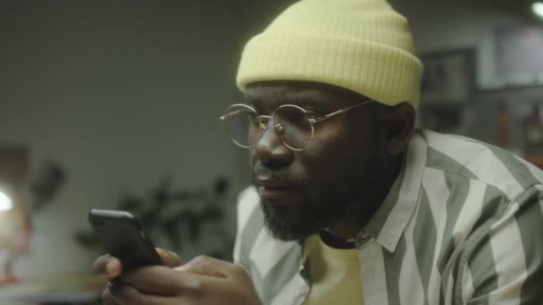 Close up shot of African American man in beanie hat and glasses wpisując wiadomość przez telefon podczas pracy wieczorem w studio - Materiał filmowy, wideo