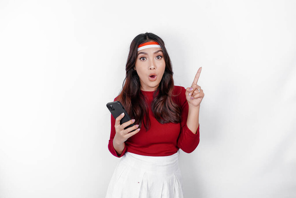 A portrait of a shocked Asian woman wearing headband, holding her phone, and pointing copy space on top of her, isolated by white background. Indonesia's independence day concept - Foto, Imagen