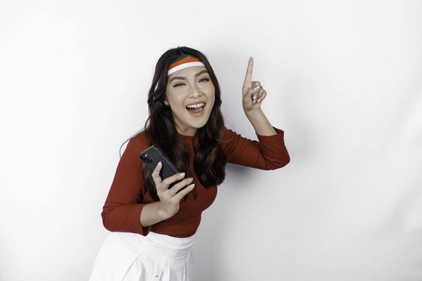 A happy Asian woman wearing headband, holding her phone, and pointing copy space on top of her, isolated by white background. Indonesia's independence day - Zdjęcie, obraz