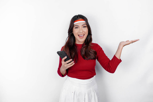 A happy Asian woman wearing headband, holding her phone, and pointing copy space beside her, isolated by white background. Indonesia's independence day - Φωτογραφία, εικόνα