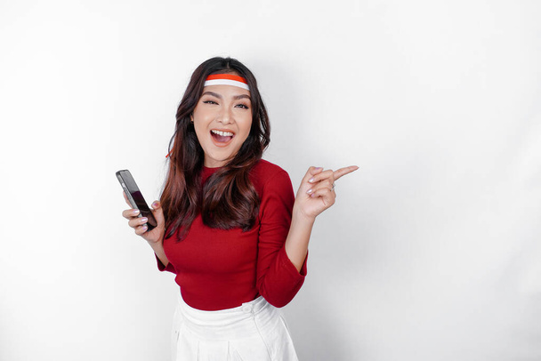 A happy Asian woman wearing headband, holding her phone, and pointing copy space beside her, isolated by white background. Indonesia's independence day - Φωτογραφία, εικόνα