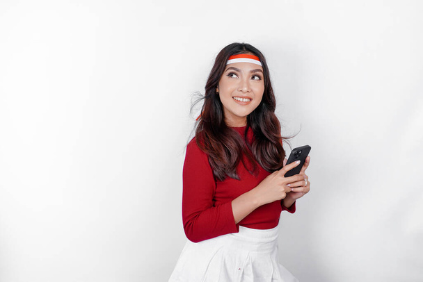 A portrait of a smiling Asian woman wearing headband and holding her phone, isolated by white background. Indonesia's independence day concept - Valokuva, kuva