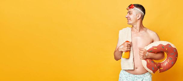 Smiling handsome adult man wearing shorts swimsuit snorkeling goggles holding donut rubber ring and bag isolated on yellow background looking at copy space standing with cream on face - Foto, immagini