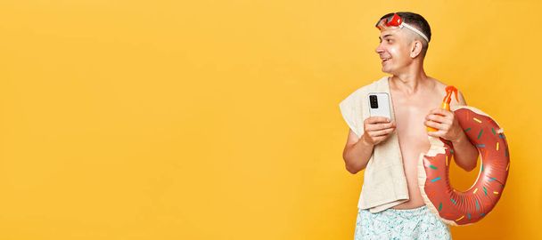 Happy cheerful adult man wearing shorts swimsuit snorkeling goggles holding donut rubber ring and bag isolated on yellow background looking away at copyspace for promotional text, resort advertisement - Valokuva, kuva