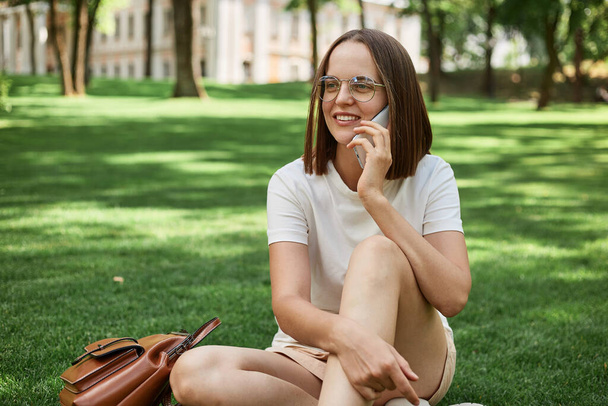 Smiling dark haired woman sitting on grass in park enjoying nature talking on mobile phone wearing casual clothing having smartphone conversation. - Zdjęcie, obraz
