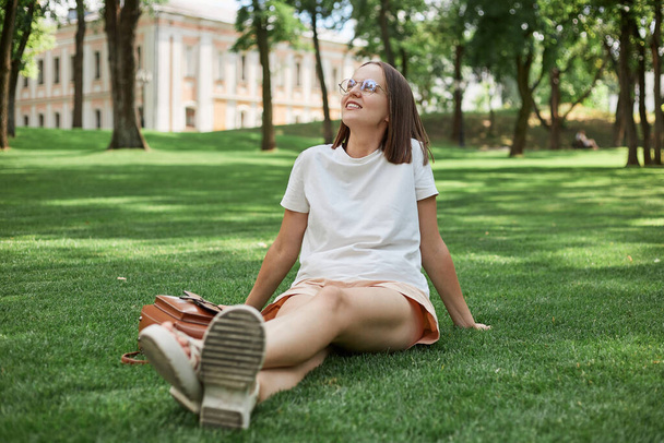 Happy young woman sitting on grass enjoining nature, carefree girl resting in city park experiencing mindfulness and calmness wearing white t shirt and shorts. - Φωτογραφία, εικόνα