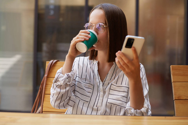 Young beautiful woman wearing striped blouse and eyeglasses drinking coffee from paper cup and looking away while holding smartphone sitting at cafeteria. - 写真・画像