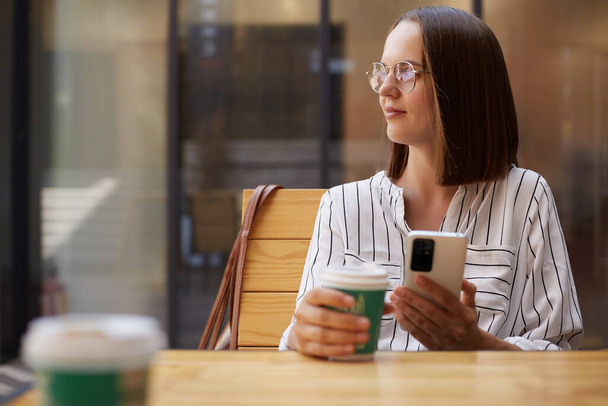 Молода приваблива жінка тримає каву в паперовій чашці і дивиться в сторону за допомогою смартфона, сидячи в кафе в смугастій блузці та окулярах. - Фото, зображення