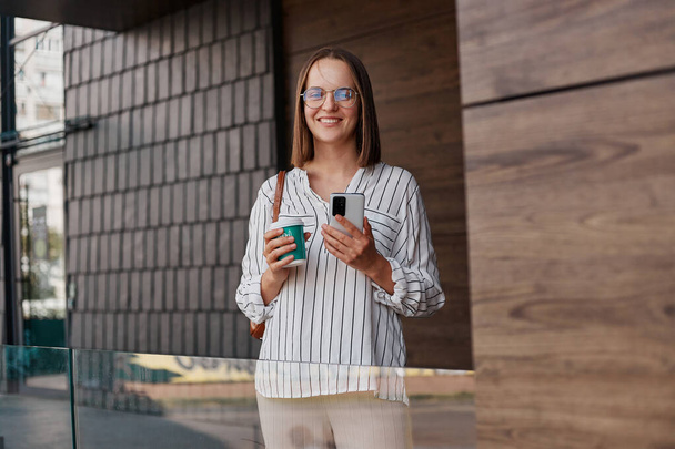 Outdoor shot van Kaukasische glimlachende aantrekkelijke vrouw dragen elegante kleding staan op straat in de buurt van het gebouw drinken van afhaalmaaltijden koffie en met behulp van mobiele telefoon chatten online. - Foto, afbeelding