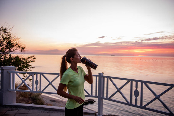 Portrait of an athlete who drinks water from a sports bottle. A young woman quenches her thirst with a fitness drinker. Maintaining water balance during training at sunrise. Girl's morning routine. - Foto, afbeelding