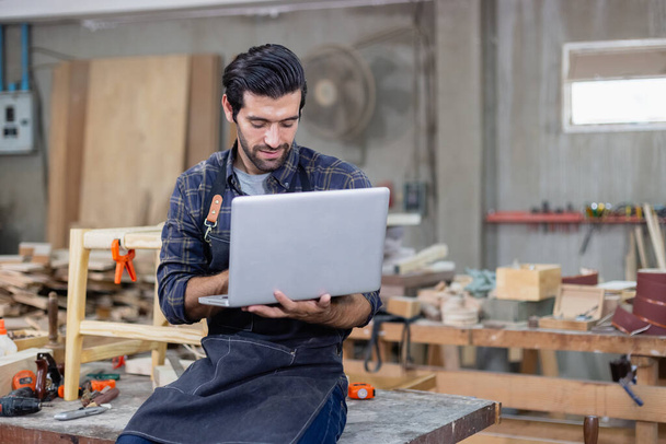 young carpenter caucasian man with laptop make drawing on wood to design at workspace. craftsman profession in wood factory - Φωτογραφία, εικόνα