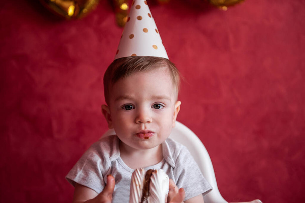 Little boy of one year old in party hat sits in childrens chair, eating birthday cake, Meringue, blows out candle. Kid on red isolated background, on textured wall foil gold balloons One. Home party - Photo, image