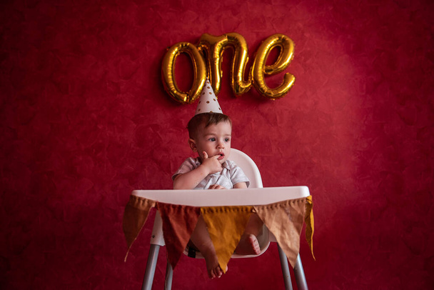 Portrait of Little boy of one year in festive hat sits in childrens chair with fabric flags. Kid on red isolated background with foil gold balloons One, on textured wall celebrating holiday at home - Foto, afbeelding