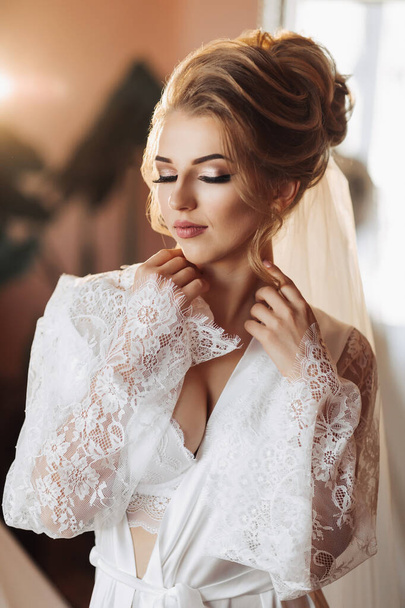 A blonde bride in a long, lacy petticoat is standing in her room, posing next to her wedding dress. Beautiful hair and make-up, open bust - Fotografie, Obrázek