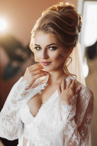 長いレースのペチコートを着たブロンドの花嫁が彼女の部屋に立っていて、結婚式のドレスの横にポーズをとっている。美しい髪と化粧、オープンバスト - 写真・画像