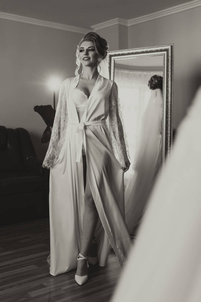 Black and white photo. A blonde bride in a long, lacy petticoat is standing in her room, posing by the mirror, putting on her earrings. Beautiful hair and makeup, open bust. Wedding portrait. Reflection in the mirror - Фото, изображение