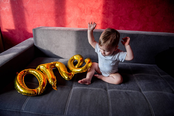 First birthday party, baby plays with gold colored foil balloons ONE. Little boy fooling around, sitting on gray sofa in sun. Light shadow drawing on red wall. Atmospheric lifestyle holiday at home - Foto, Imagem
