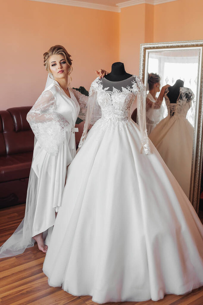 A blonde bride in a long lace robe and white shoes stands in her room next to her wedding dress, posing by the mirror. Beautiful hair and make-up, open bust. Wedding portrait. Open legs - Φωτογραφία, εικόνα