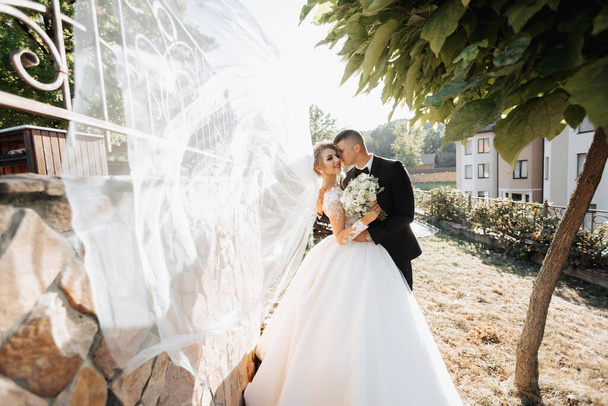 Wedding portrait. A groom in a black suit and a blonde bride stand embracing by a stone wall under a tree. A white, long veil in the air. Photo session in nature. Beautiful hair and makeup - Foto, immagini