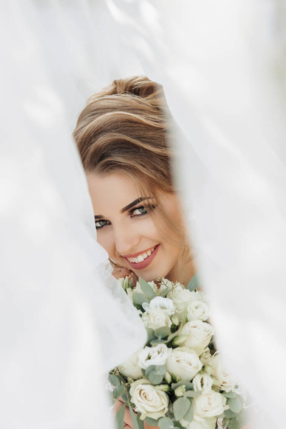 Curly blonde bride in a white dress, covered with a veil, poses for the camera with a bouquet of roses. Portrait of the bride. Beautiful makeup and hair. Wedding in nature - Fotografie, Obrázek