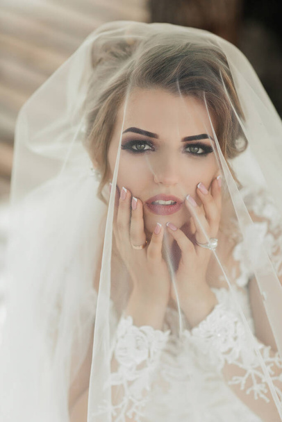 Curly blonde bride in a white dress, covered with a veil, poses for the camera with a bouquet of roses. Portrait of the bride. Beautiful makeup and hair. Wedding in nature - Foto, imagen