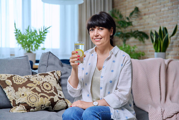 Smiling mature woman relaxing at home with glass of lemon water. 50s positive female sitting on couch in living room looking at camera. Age people, lifestyle health beauty lifestyle diet food concept - Valokuva, kuva